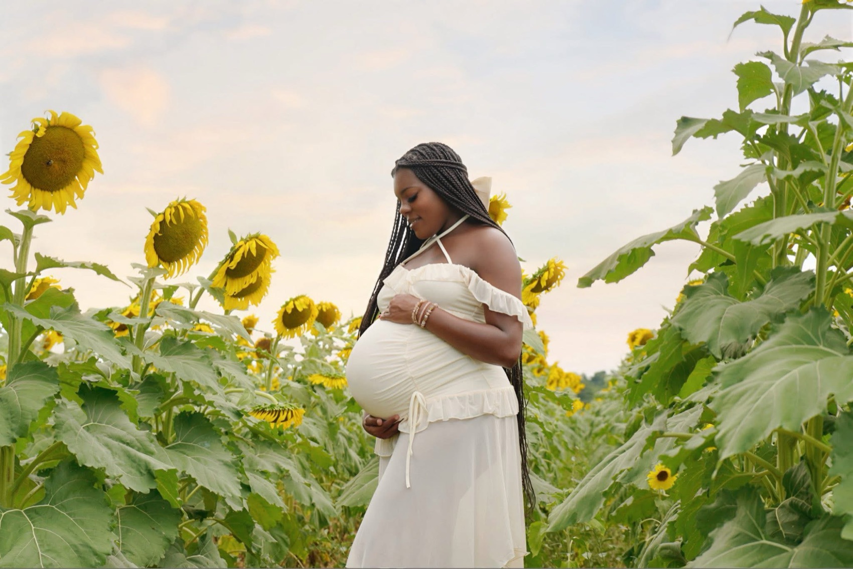 Expectant mother in a cream gown cradling her belly in a sunflower field at dusk