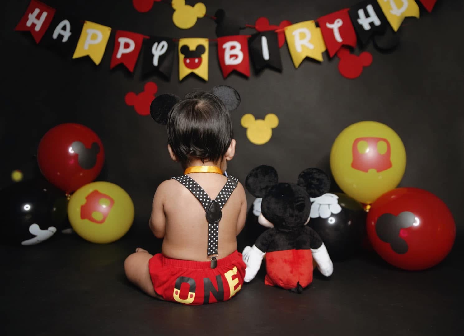 First birthday studio session: a baby in Mickey Mouse ears and red bloomers lettered ONE, sitting with a plush Mickey beneath a Happy Birthday banner and balloons
