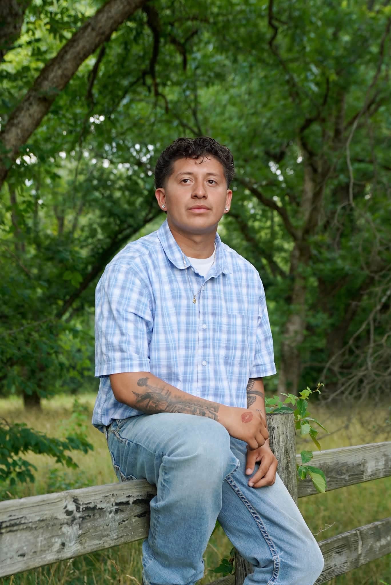 Young man in a light blue plaid shirt leaning back against a weathered split-rail fence with summer woods behind him