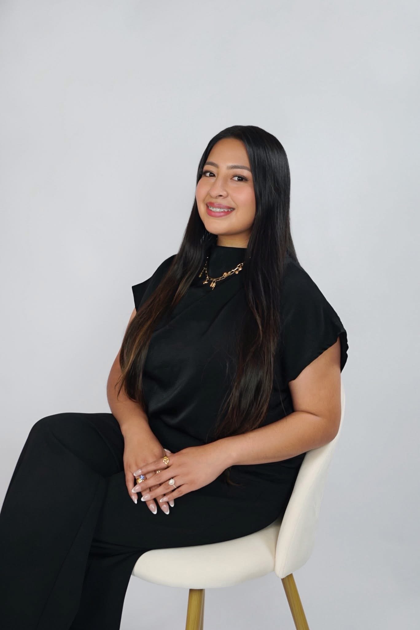 Studio portrait of a woman in a black top and trousers seated on a cream chair against a soft white backdrop