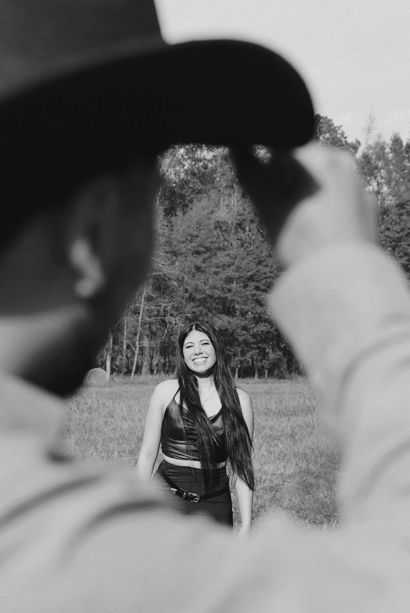 Black and white photograph framed past a man in a wide-brim hat, his partner laughing in the sunlit field beyond