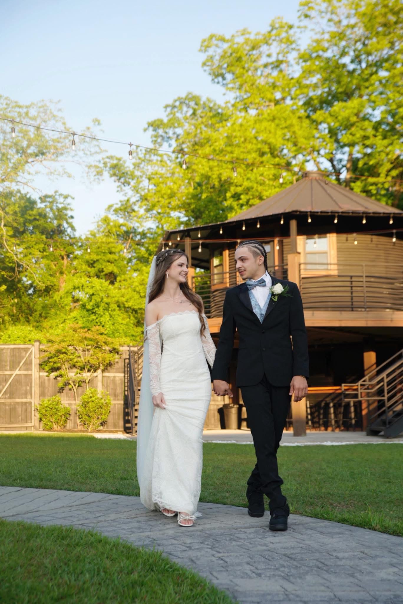 Bride in a lace off-the-shoulder gown and veil walking hand in hand with her groom along a garden path under caf&eacute; string lights